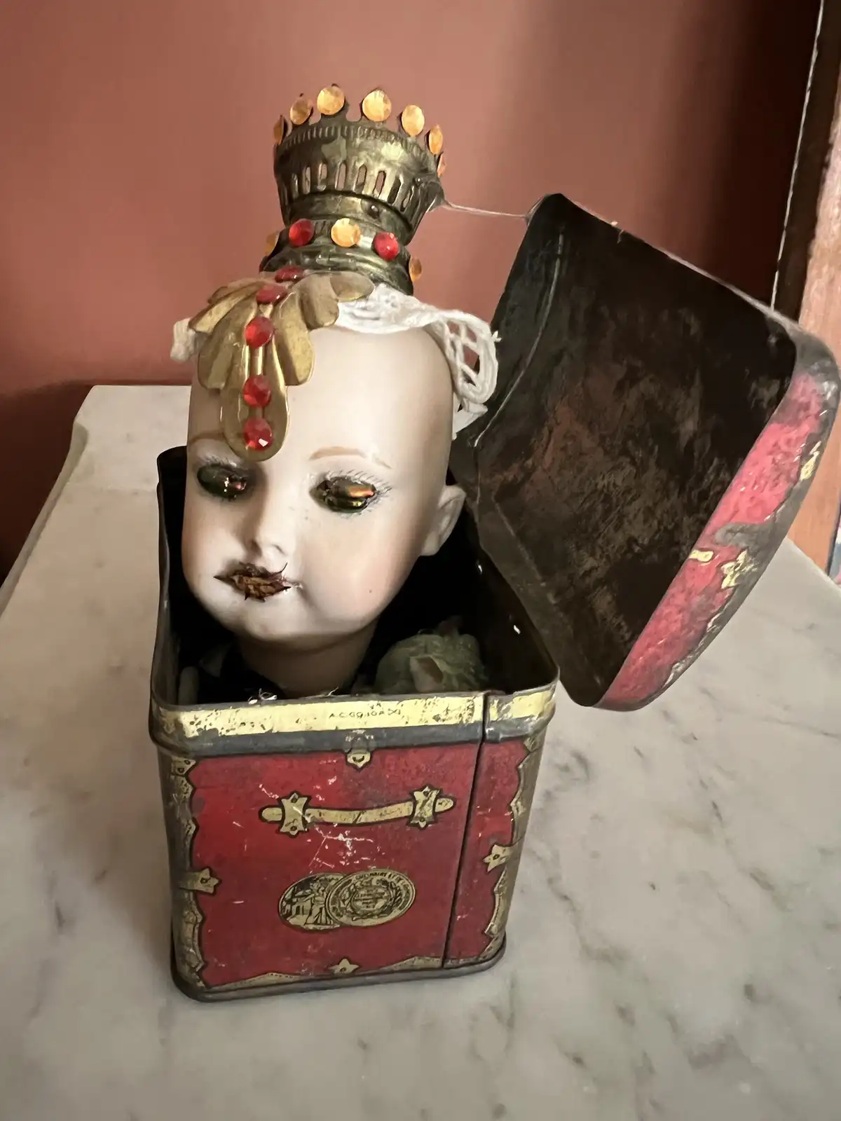 A vintage porcelain doll head with a gold crown and red jewels is emerging from an ornate, red and gold tin box. The doll…