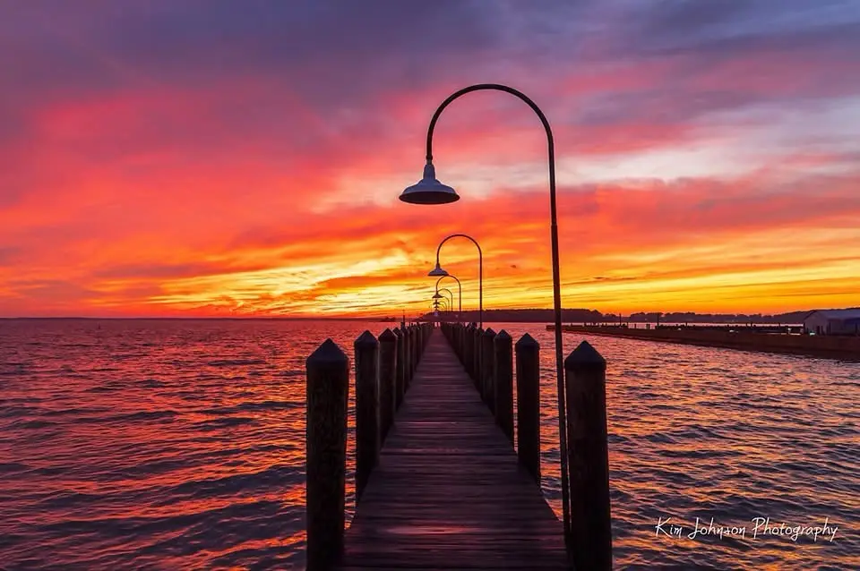 A serene sunset photograph shows a wooden pier extending into water, lined with curved lampposts silhouetted against a…