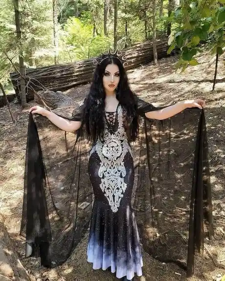 A woman with long black hair and dramatic makeup stands in a forest, wearing an ornate black and white gown with sheer black…