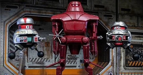 Three robot figures stand in a sci-fi setting: a large red central robot flanked by two smaller silver robots with…