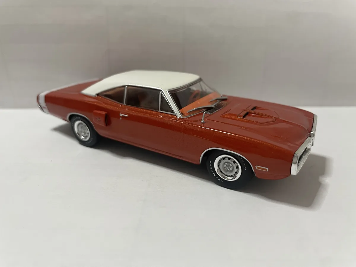 A red and white die-cast model of a classic American muscle car, likely a Dodge Charger, is photographed on a plain white…