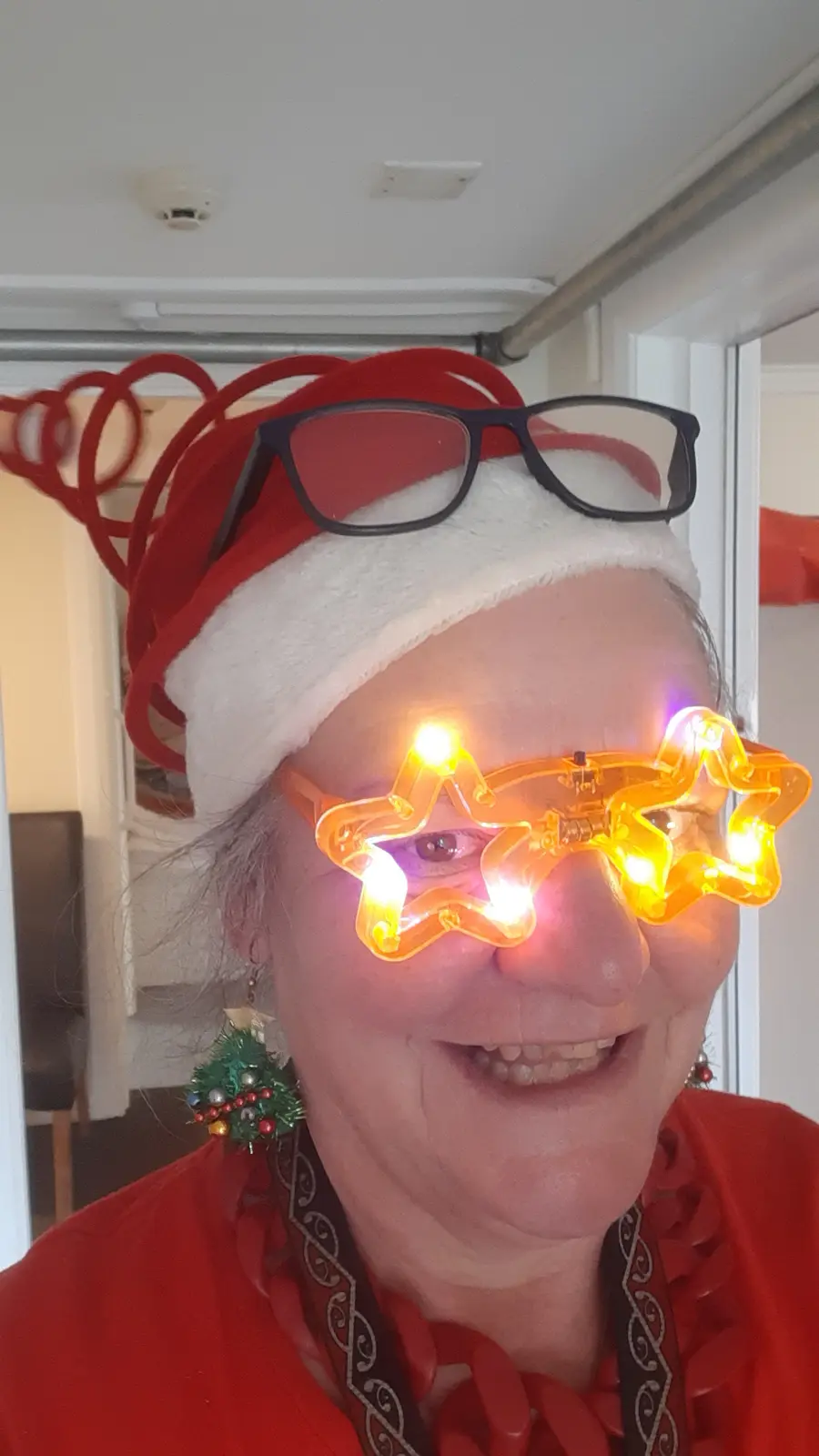 A smiling woman wears a red and white Santa hat with red spiral antlers, black-rimmed glasses perched on top, and bright…
