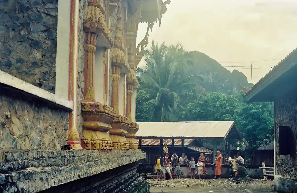 An old photograph shows a group of people, including several monks in orange robes, gathered near a traditional temple with…