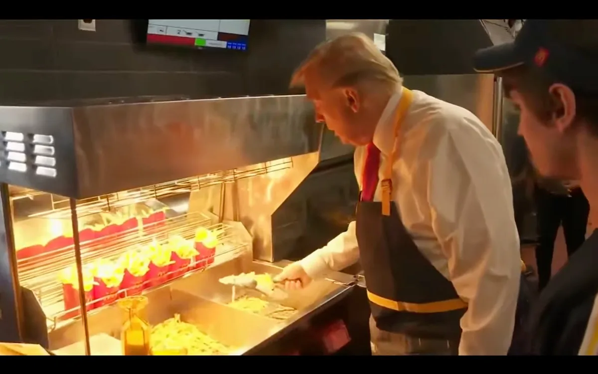 A man with distinctive blonde hair, wearing a white shirt, red tie, and black apron with yellow trim, is serving french…