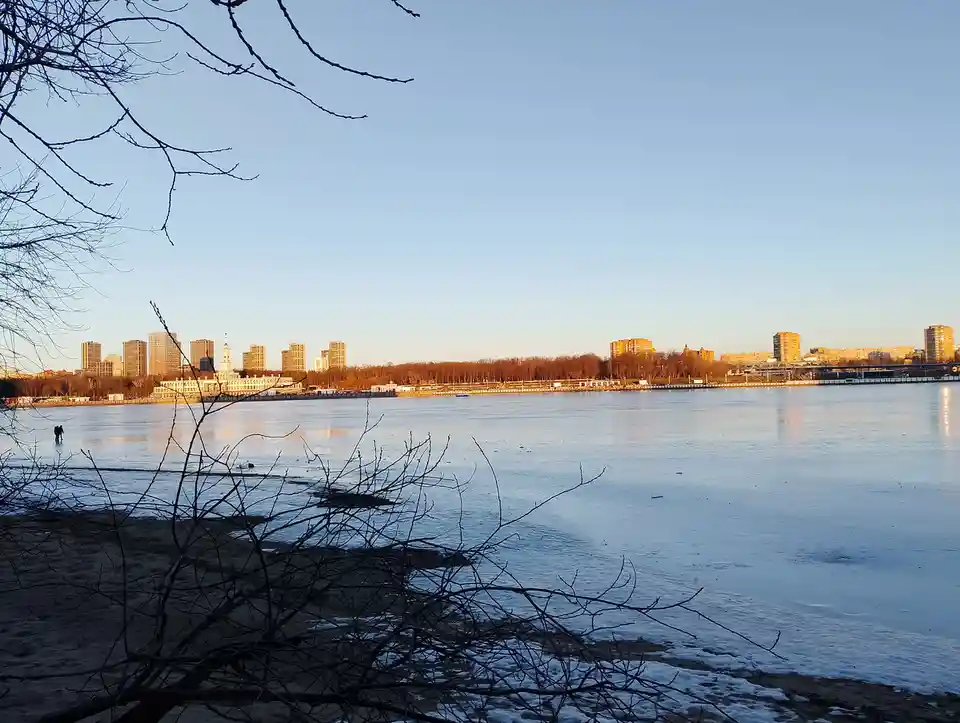 A wide shot of a frozen river with a city skyline in the background under a clear blue sky. Bare tree branches frame the…