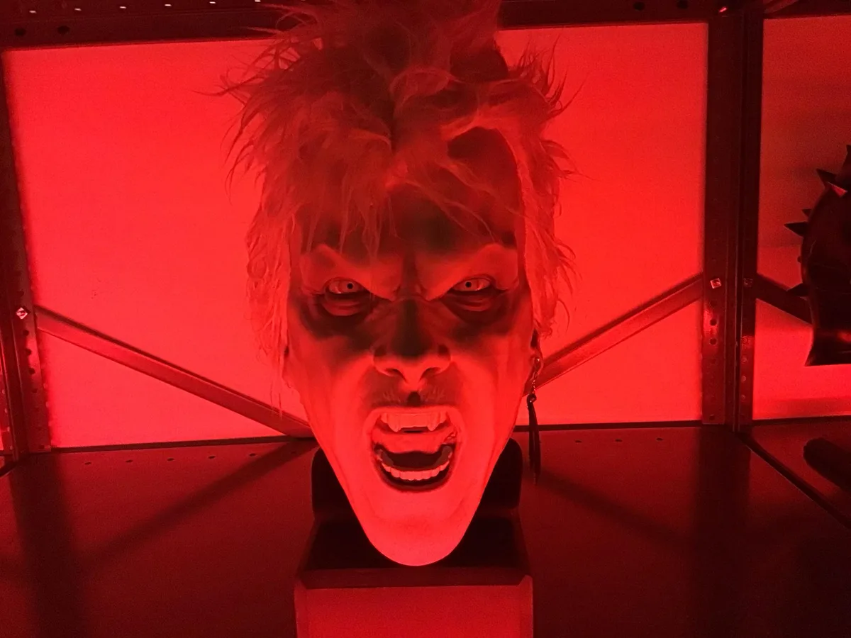 A close-up of a realistic, screaming vampire mask with spiky hair and fangs, illuminated by intense red lighting inside a…