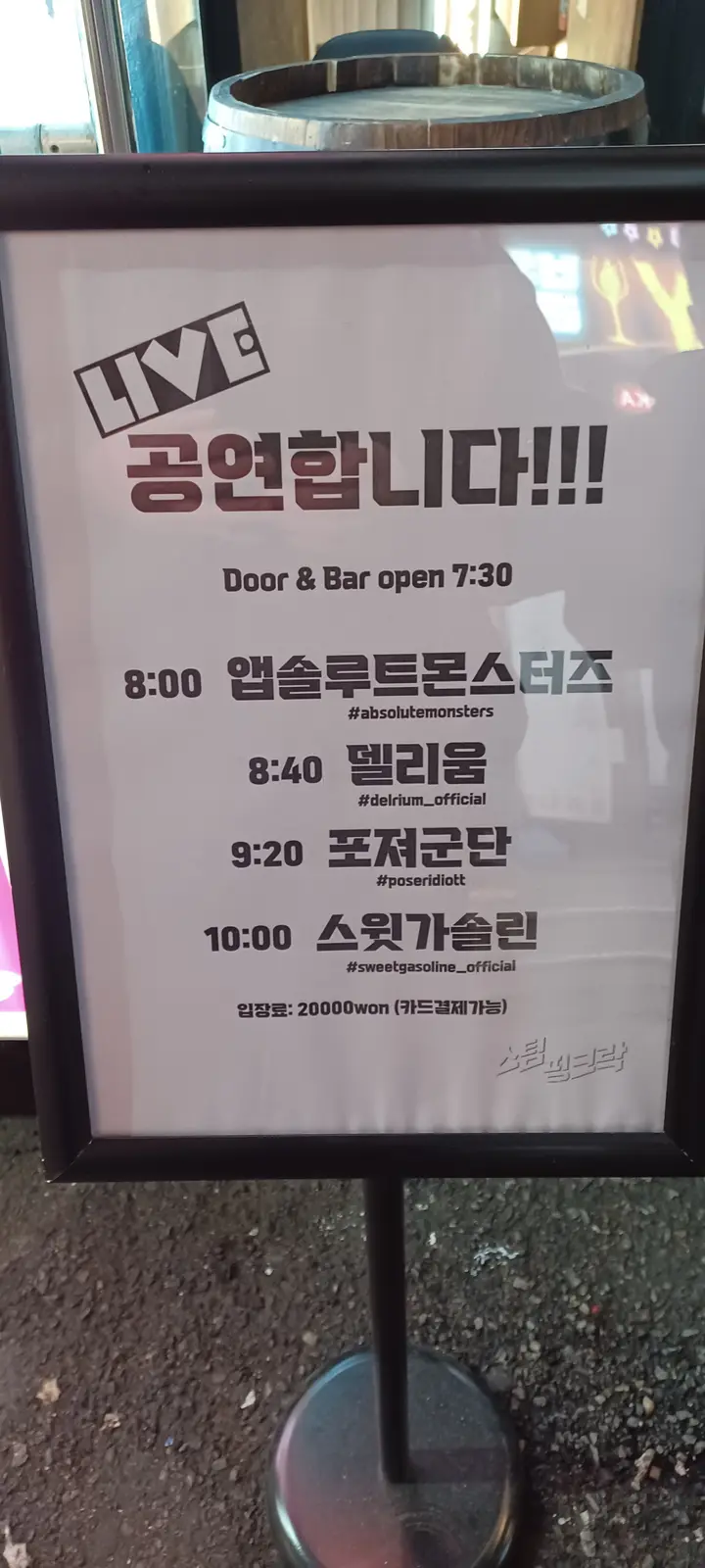 A framed poster for a live music event in Korean, listing performers and times, with 'LIVE 공연합니다!!!' at the top and a…