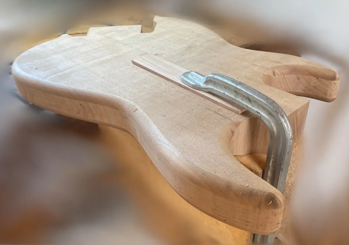 A close-up photo shows a light-colored wooden guitar body being held in a metal clamp, with a small wooden strip placed…