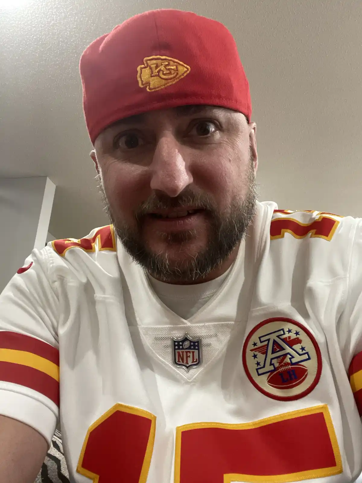 a man with a beard and wide eyes wears a red kansas city chiefs cap and a white jersey with red and yellow accents. The…