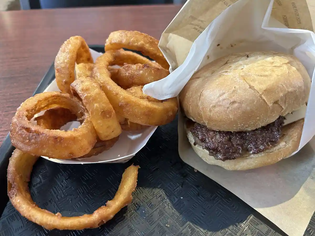 A close-up photo shows a burger in a paper wrapper and a side of onion rings on a black tray. The burger appears to be just…