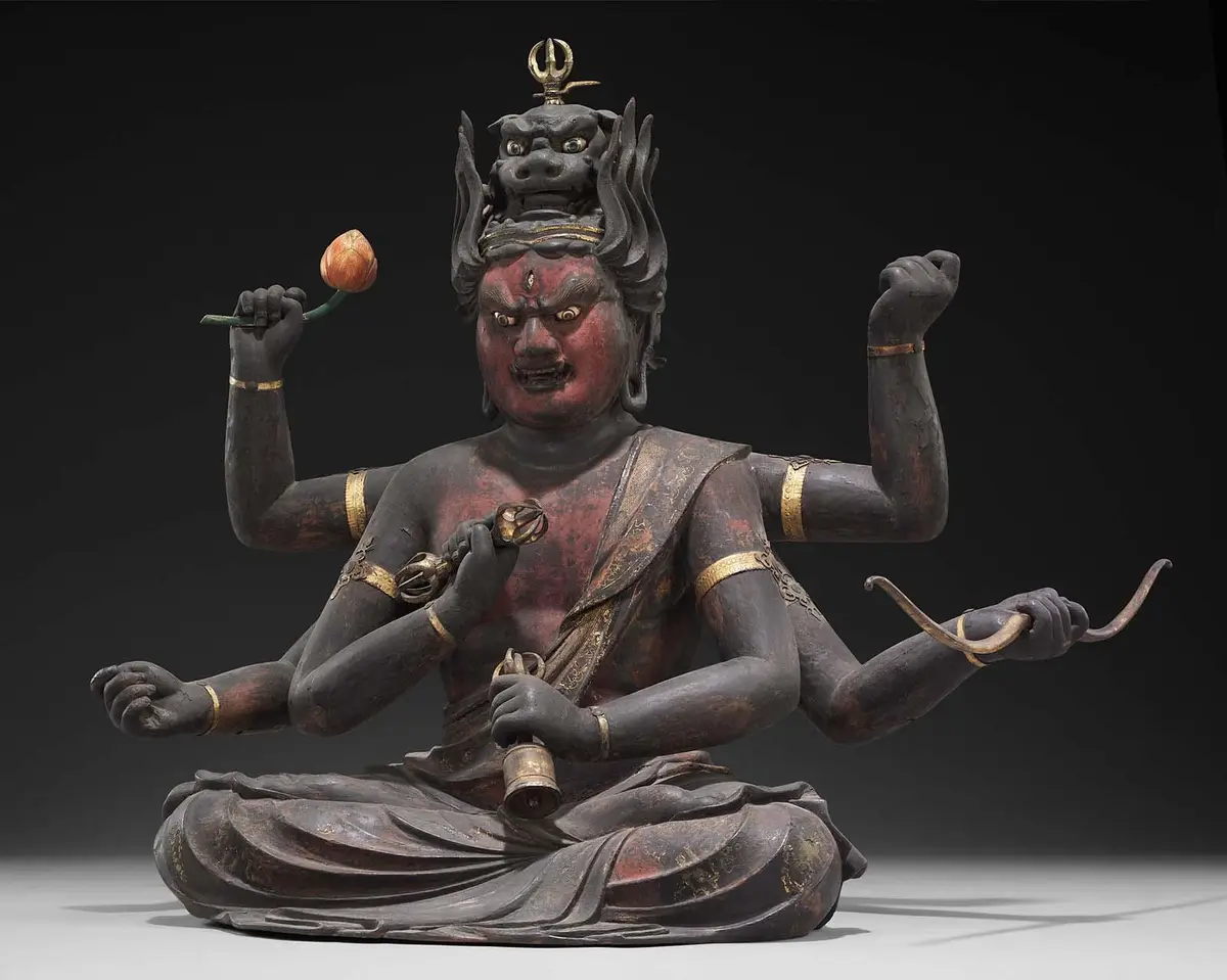 A dark-colored, multi-armed statue of a wrathful Buddhist deity with a fierce red face, holding a lotus, a bell, and a…