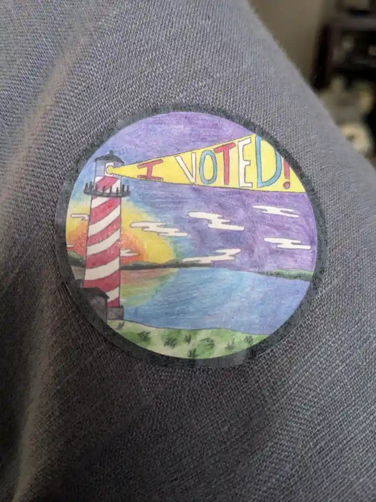 A hand-drawn, circular 'I VOTED!' sticker is pinned to a gray fabric surface. The sticker depicts a red-and-white striped…