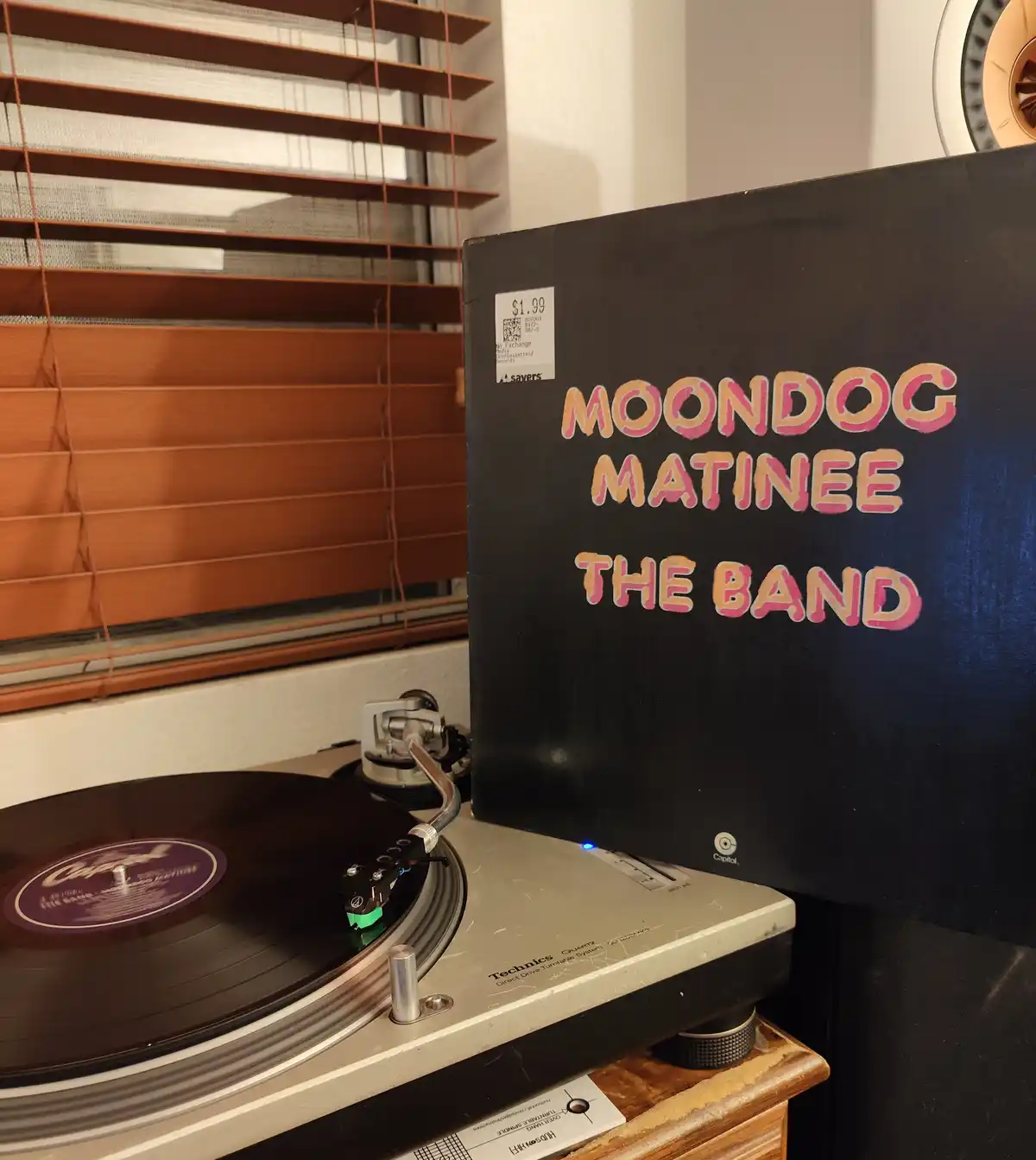 A vinyl record sleeve for 'Moondog Matinee The Band' leans against a Technics turntable with a record spinning. The sleeve…