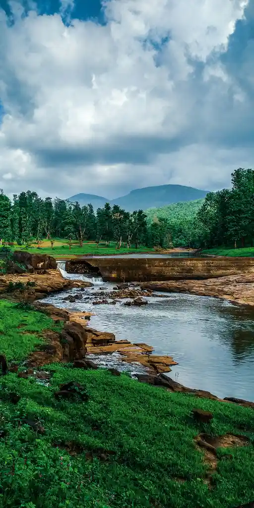 A scenic landscape photo of a river flowing over rocks with a small waterfall, surrounded by lush green grass, trees, and…