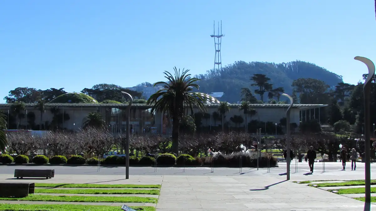 A sunny outdoor plaza with palm trees, modern curved lampposts, and a large building with green domes in the background. A…