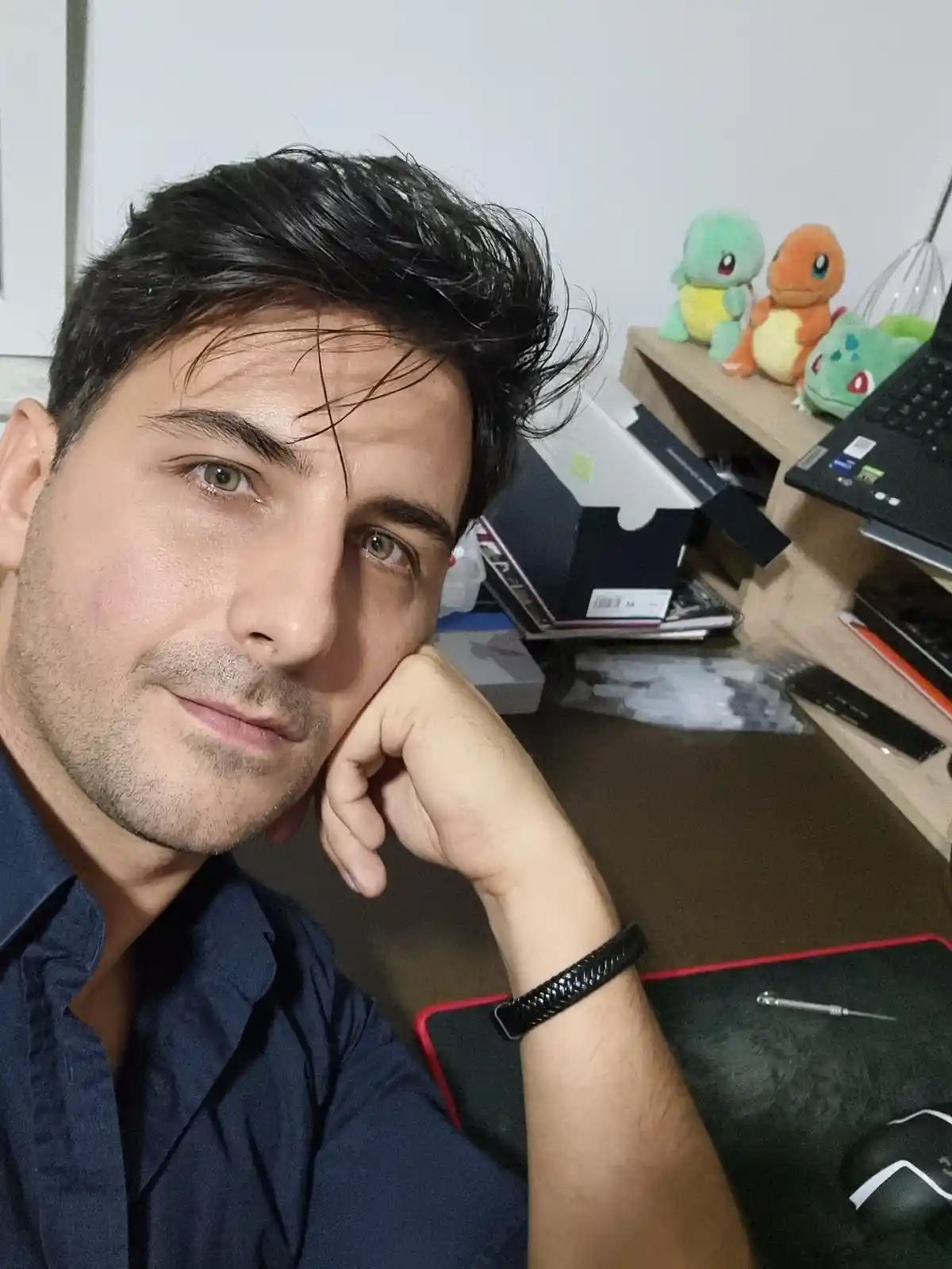 A man with dark hair and a slight beard rests his head on his hand, looking directly at the camera with a calm, slightly…