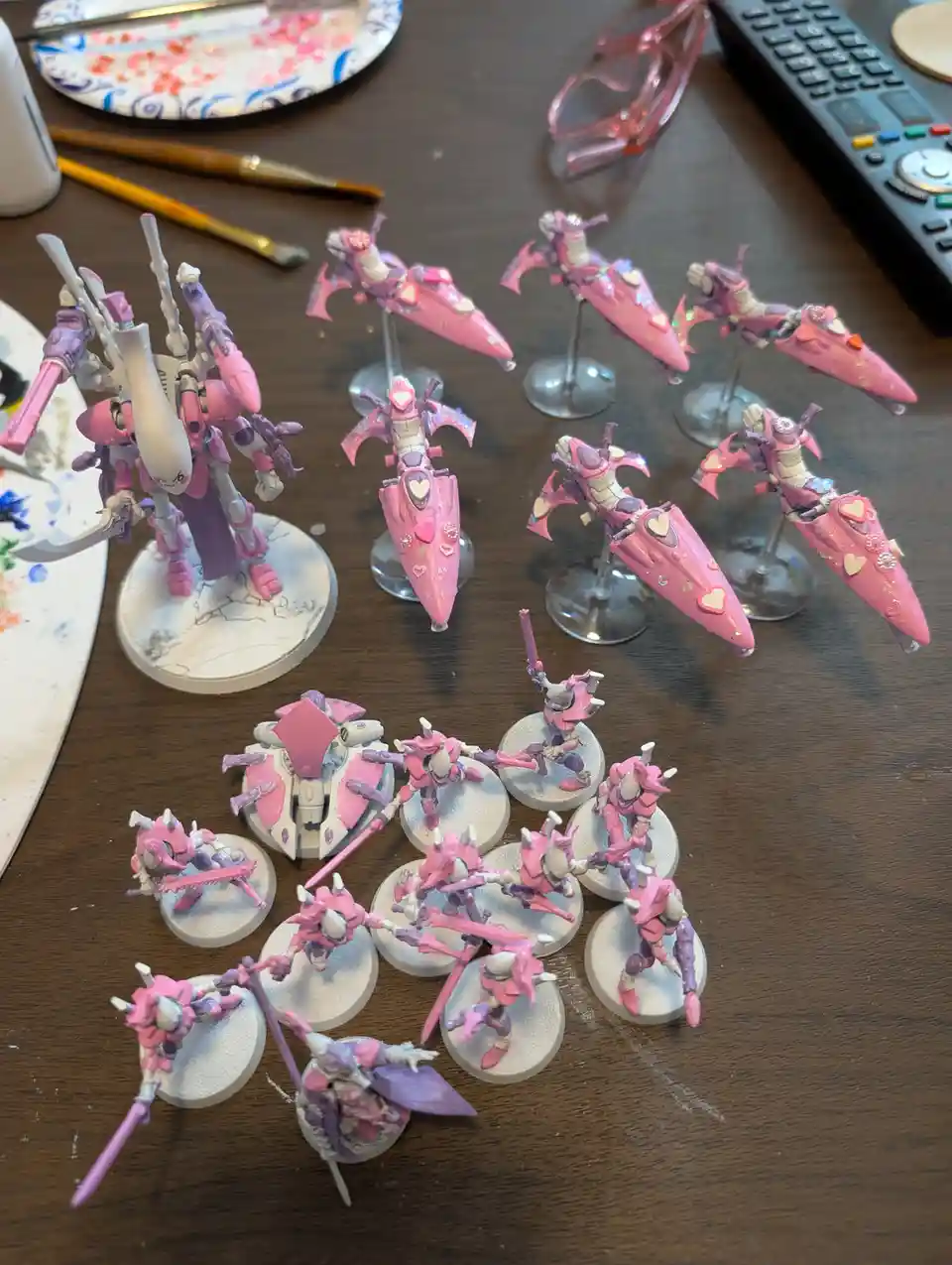 A collection of miniature sci-fi war models, mostly painted in pink with hearts and glitter, are displayed on a wooden table…