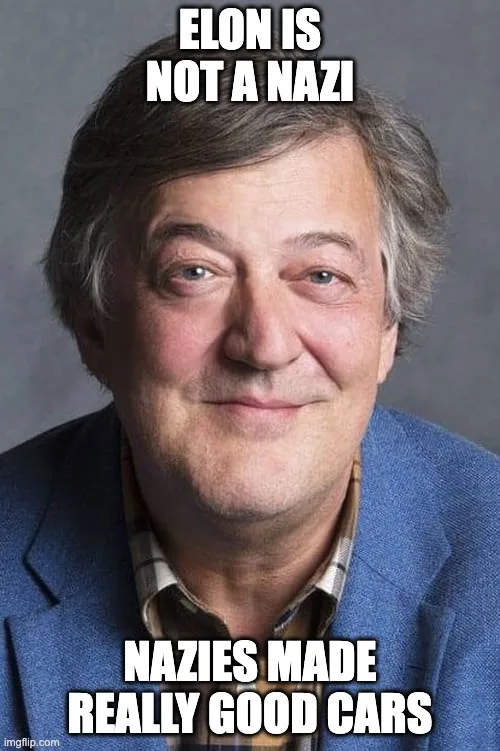 A man with graying hair, wearing a blue blazer over a plaid shirt, smiles subtly at the camera against a gray background…