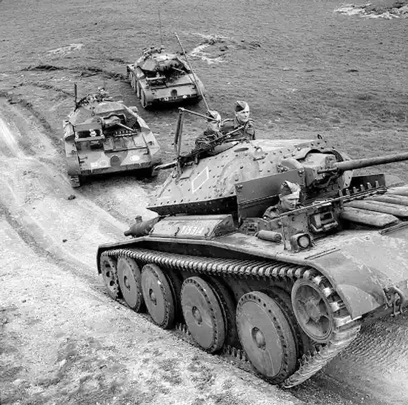 A black-and-white historical photo shows three military tanks driving in a line along a muddy dirt track. Soldiers are…