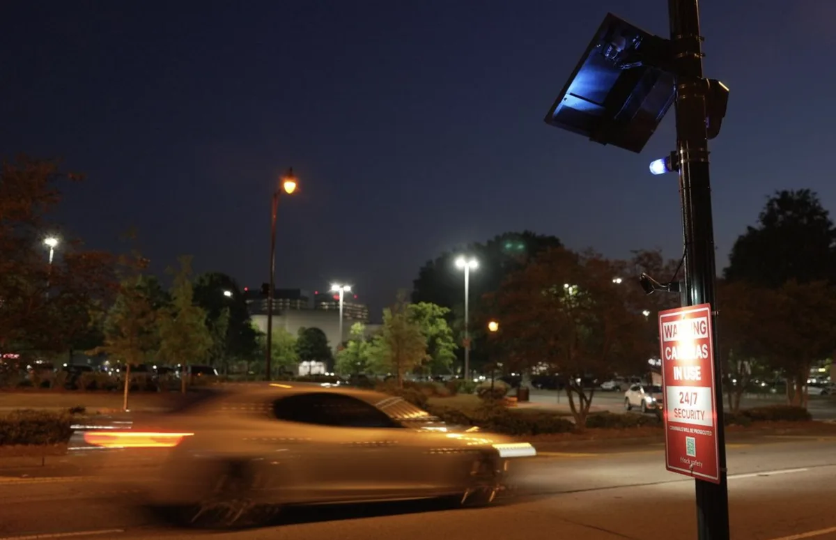 A nighttime street scene with a blurred car speeding past, next to a pole with a solar-powered security camera and a red…
