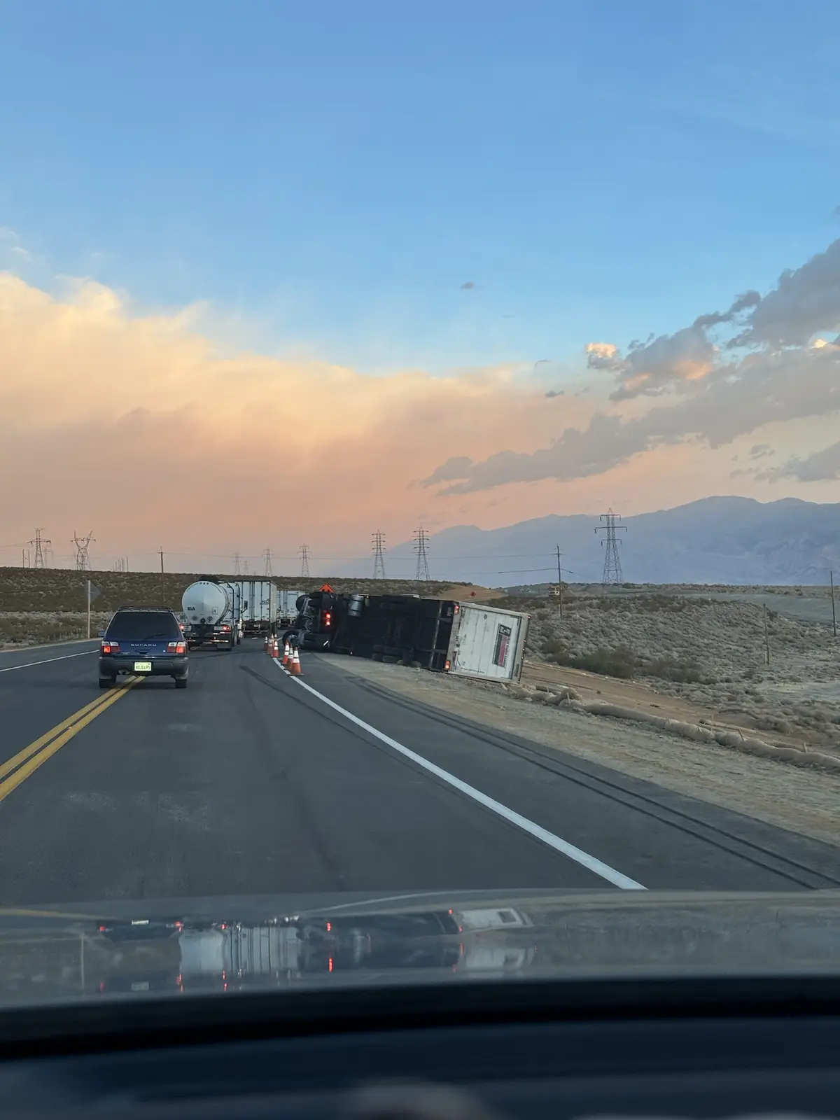 A dashcam view from a car on a highway during sunset, showing a line of stopped vehicles ahead due to a truck overturned on…