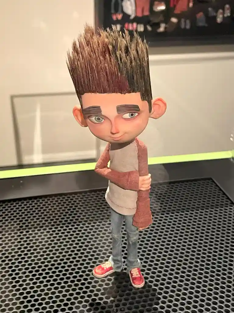 A stylized, cartoonish figurine of a young boy with spiky brown hair, thick eyebrows, and a smug smirk stands with arms…