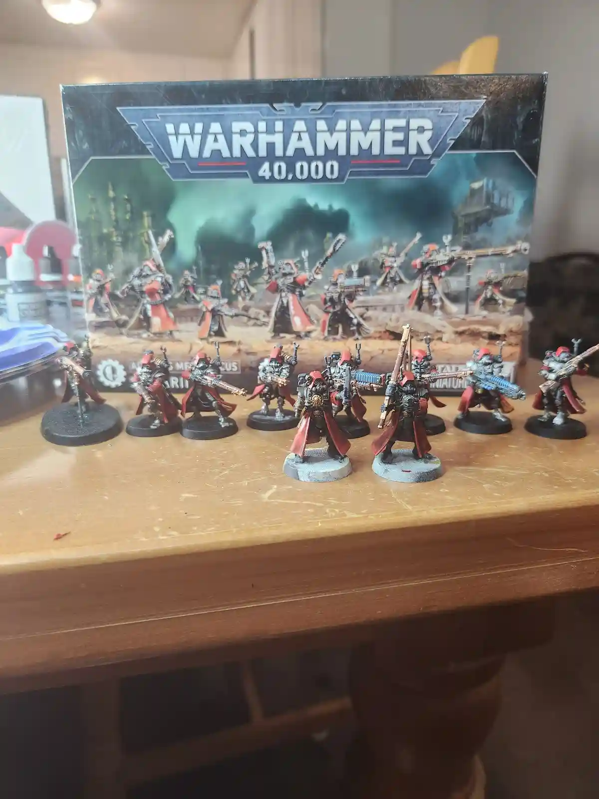 A row of painted Warhammer 40,000 miniatures, specifically Adeptus Mechanicus models in red and grey armor, are displayed on…