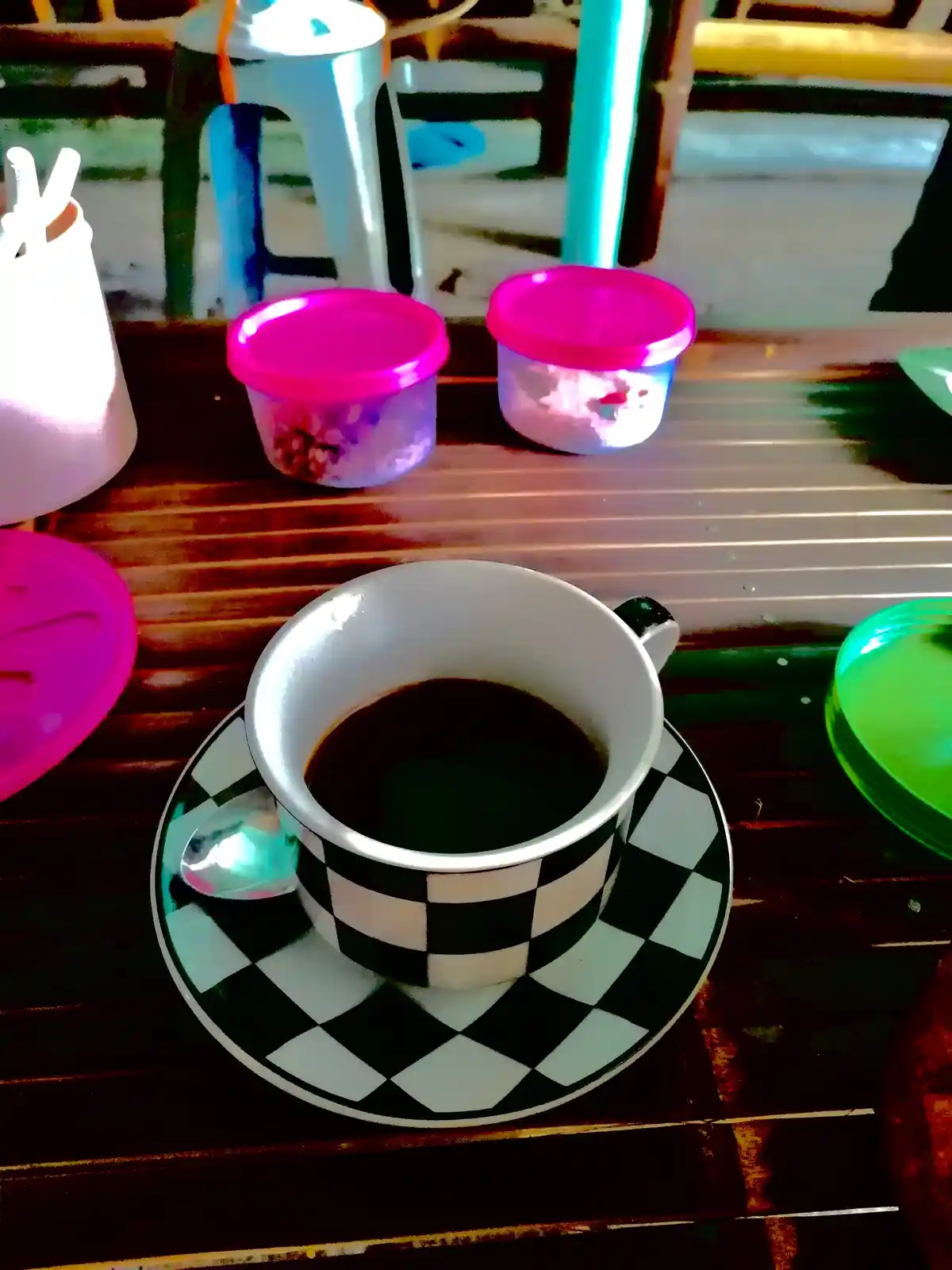 A black-and-white checkered coffee cup and saucer filled with dark coffee sits on a wooden table, surrounded by colorful…