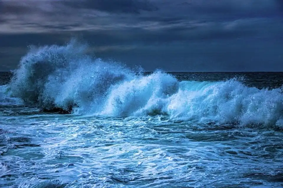 A dramatic, dark blue ocean scene with a massive, crashing wave forming a turbulent wall of white foam under a stormy sky…