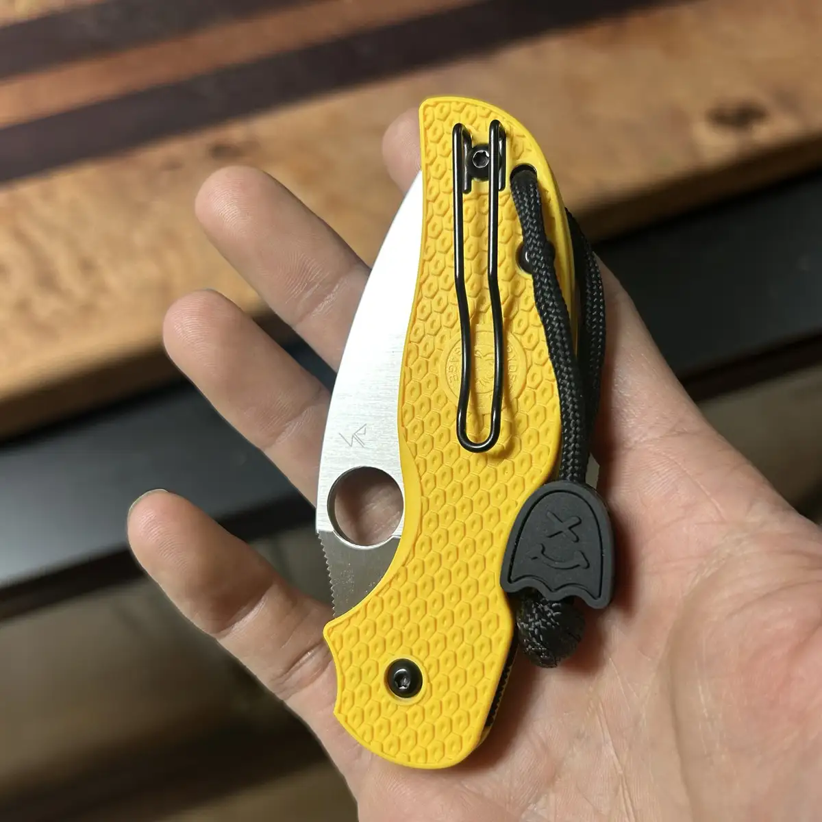 A hand holds a yellow tactical folding knife with a textured honeycomb grip, a black lanyard, and a black ghost-shaped charm…