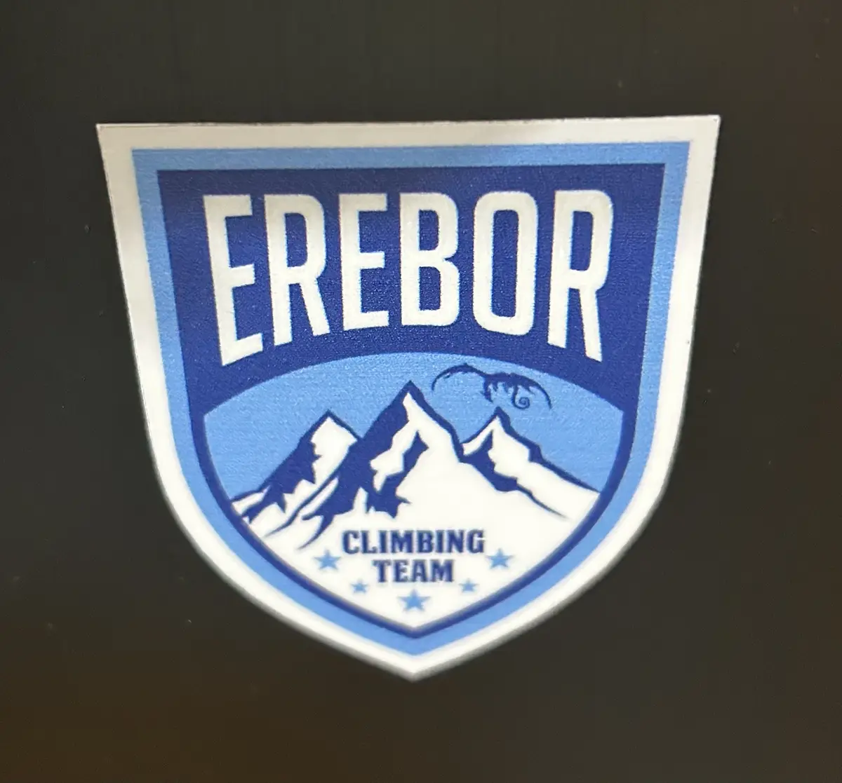 A blue and white shield-shaped patch features the word 'EREBOR' in large white letters at the top, with a stylized mountain…
