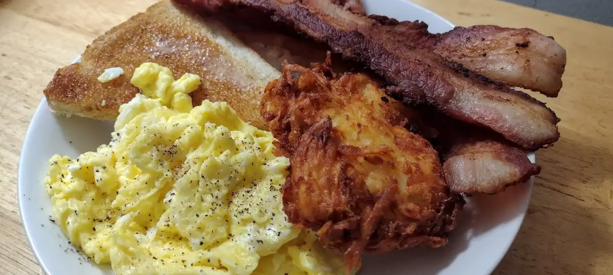 A close-up photo of a hearty breakfast plate featuring scrambled eggs seasoned with black pepper, crispy bacon strips, a…