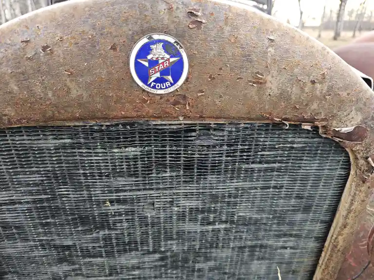 A close-up of a heavily rusted vintage car's front grille and hood, showing a faded circular 'Star Four Durant Motors'…