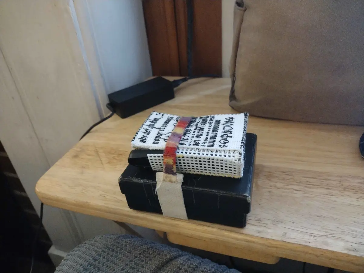 A small, handmade-looking book with a white woven cover and black text sits atop a larger black book on a wooden table. The…