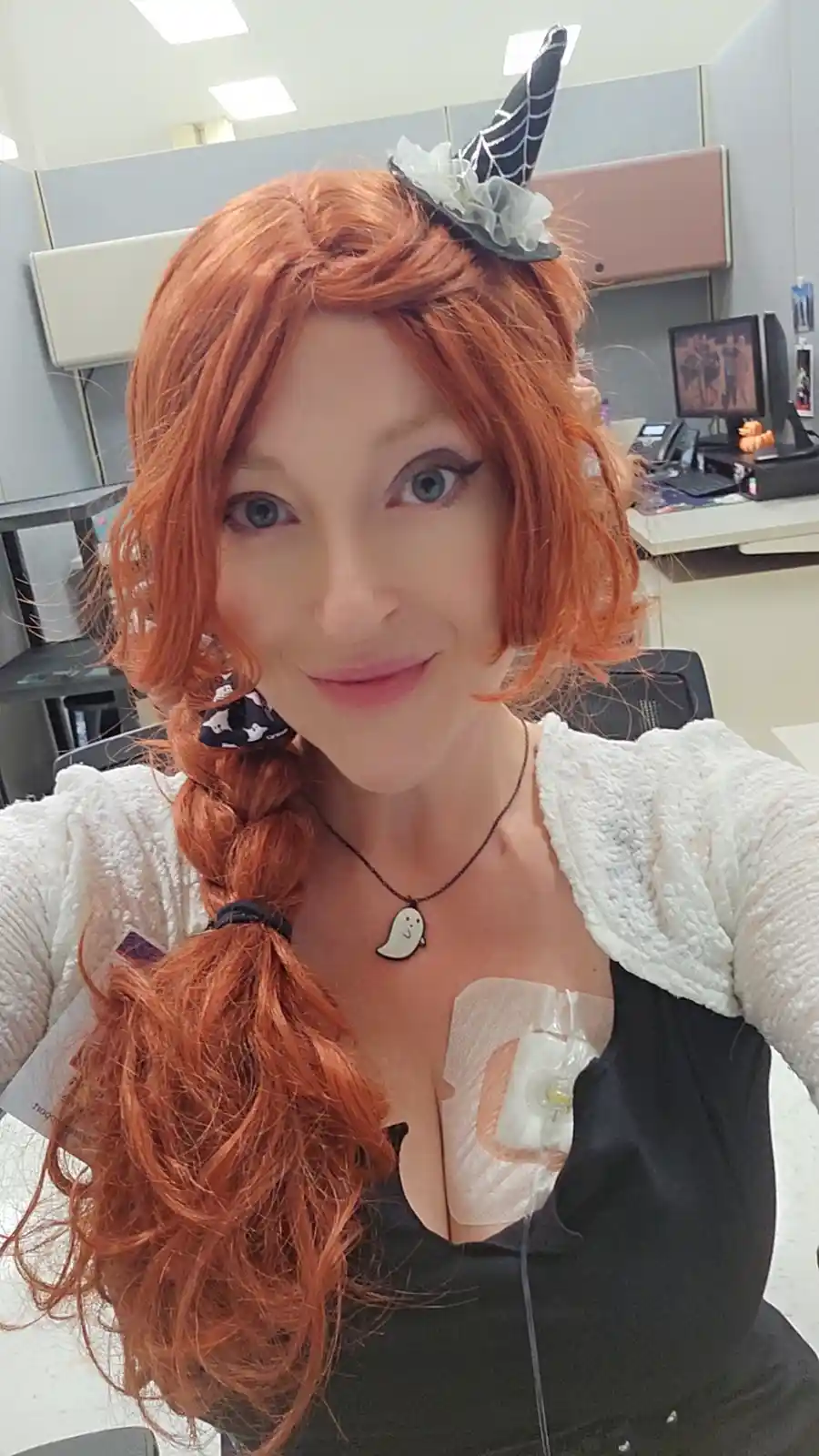 A woman with orange hair in a braid and a spider-themed headpiece smiles at the camera in an office cubicle. She wears a…