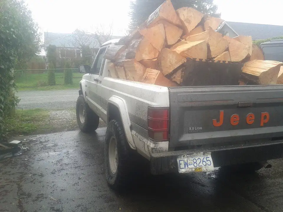 A white, older model Jeep pickup truck is parked on a wet driveway, its bed overflowing with a large pile of freshly cut…