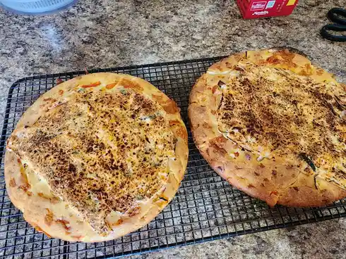 Two round, golden-brown baked pizzas sit side-by-side on a black wire cooling rack on a speckled granite countertop. The…