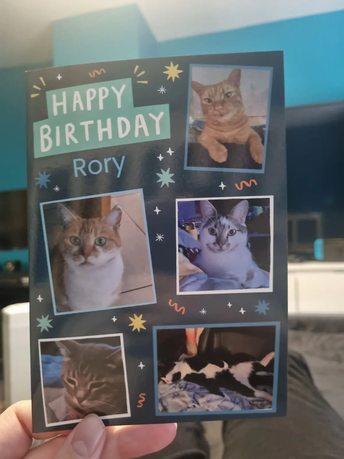 A hand holds a dark blue birthday card decorated with stars and swirls, featuring the text 'HAPPY BIRTHDAY Rory' in large…
