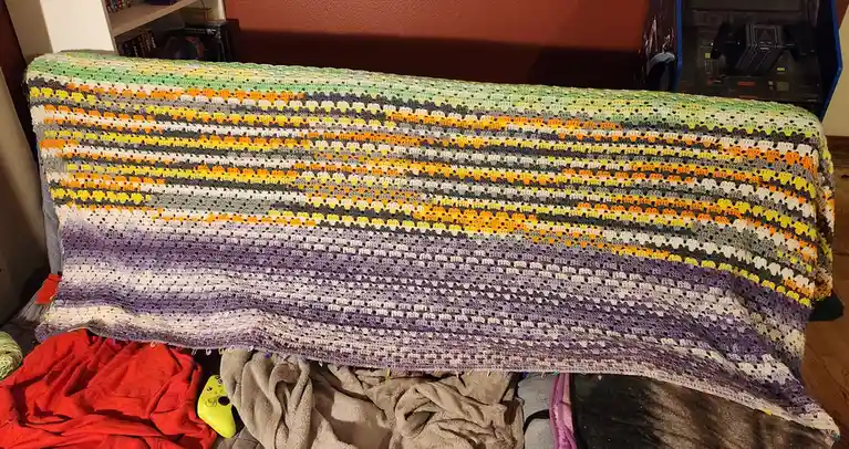 A long, hand-crocheted blanket with a gradient of colors from purple at the bottom to green and orange at the top is draped…
