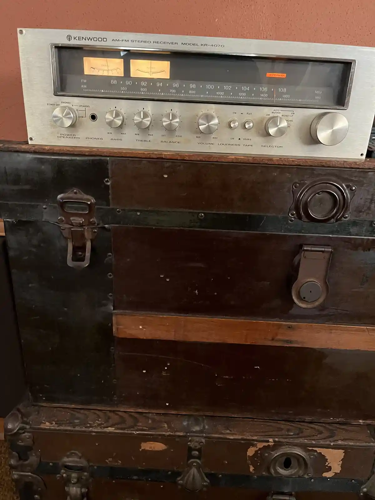 A vintage Kenwood AM-FM stereo receiver model KR-4070 sits atop a stack of three old, worn, dark brown trunks with metal…