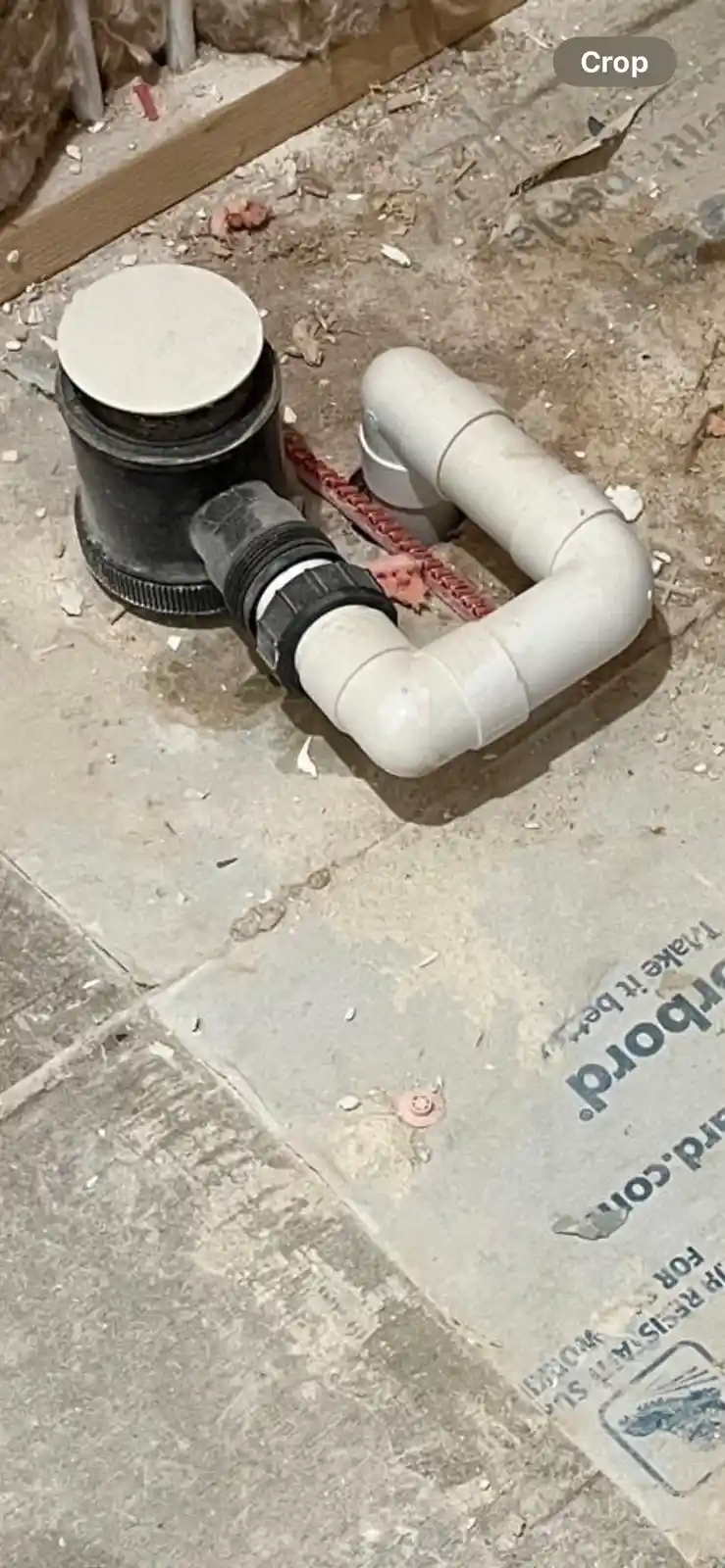 A photo shows a white PVC pipe assembly connected to a black plumbing fixture on a concrete floor, surrounded by…