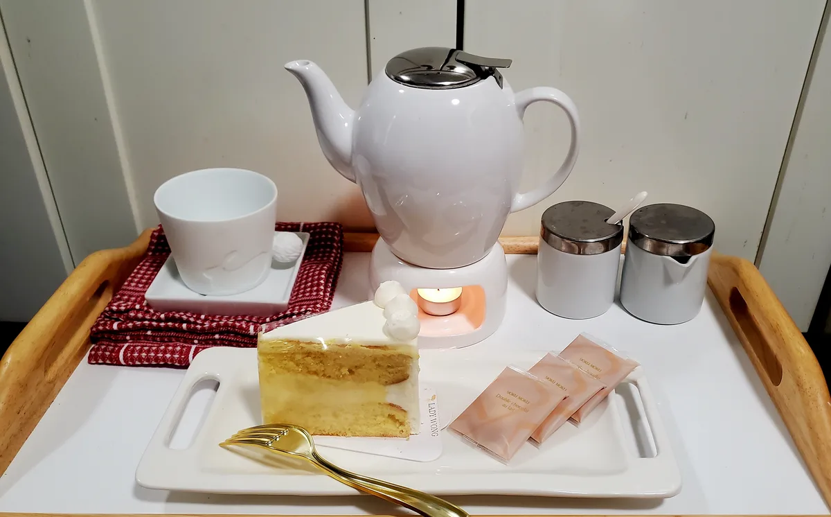 A wooden tray holds a white teapot on a warmer with a lit candle, a white teacup on a red cloth, a slice of layered cake…