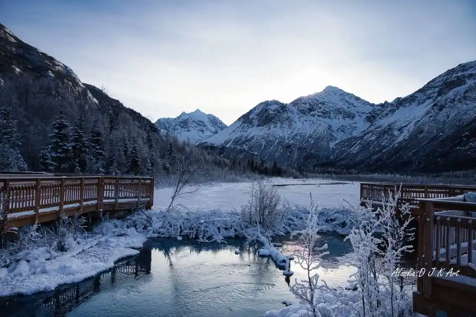 A serene winter landscape photograph shows snow-covered mountains, a partially frozen river, and wooden boardwalks with snow…