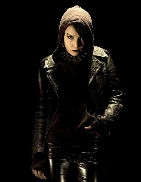 A person with dark makeup and a serious expression wears a dark hoodie under a black leather jacket with silver accents…