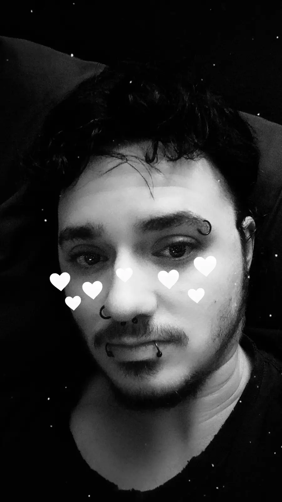A black-and-white selfie of a man with multiple facial piercings, including a nose ring and lip piercing, lying down with a…