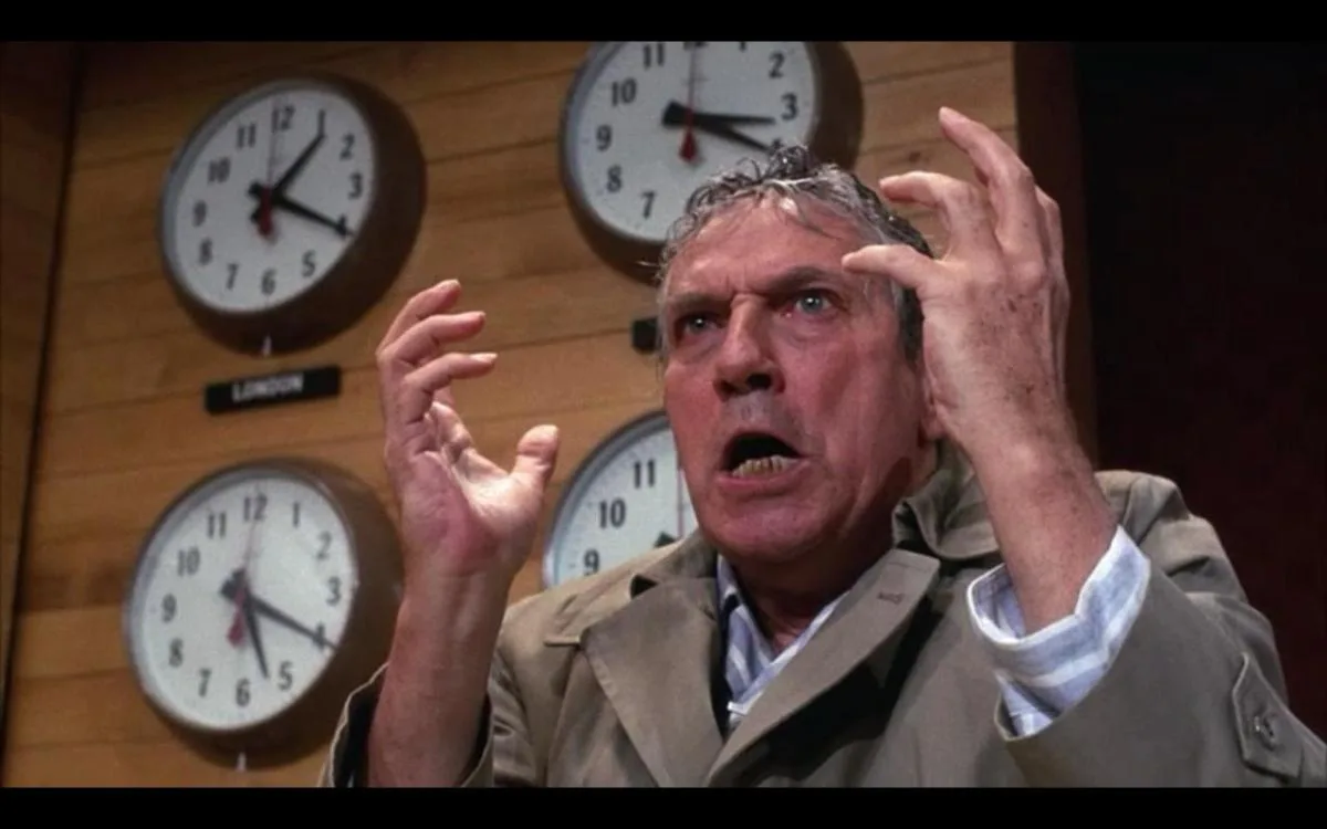 a man in a trench coat stands before a wall of clocks, his hands raised in panic as he stares upward with a horrified…