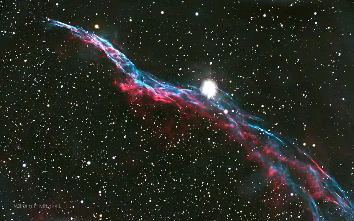 A cosmic image of a vibrant red and blue nebula streaking across a star-filled black sky, with a bright star near its…