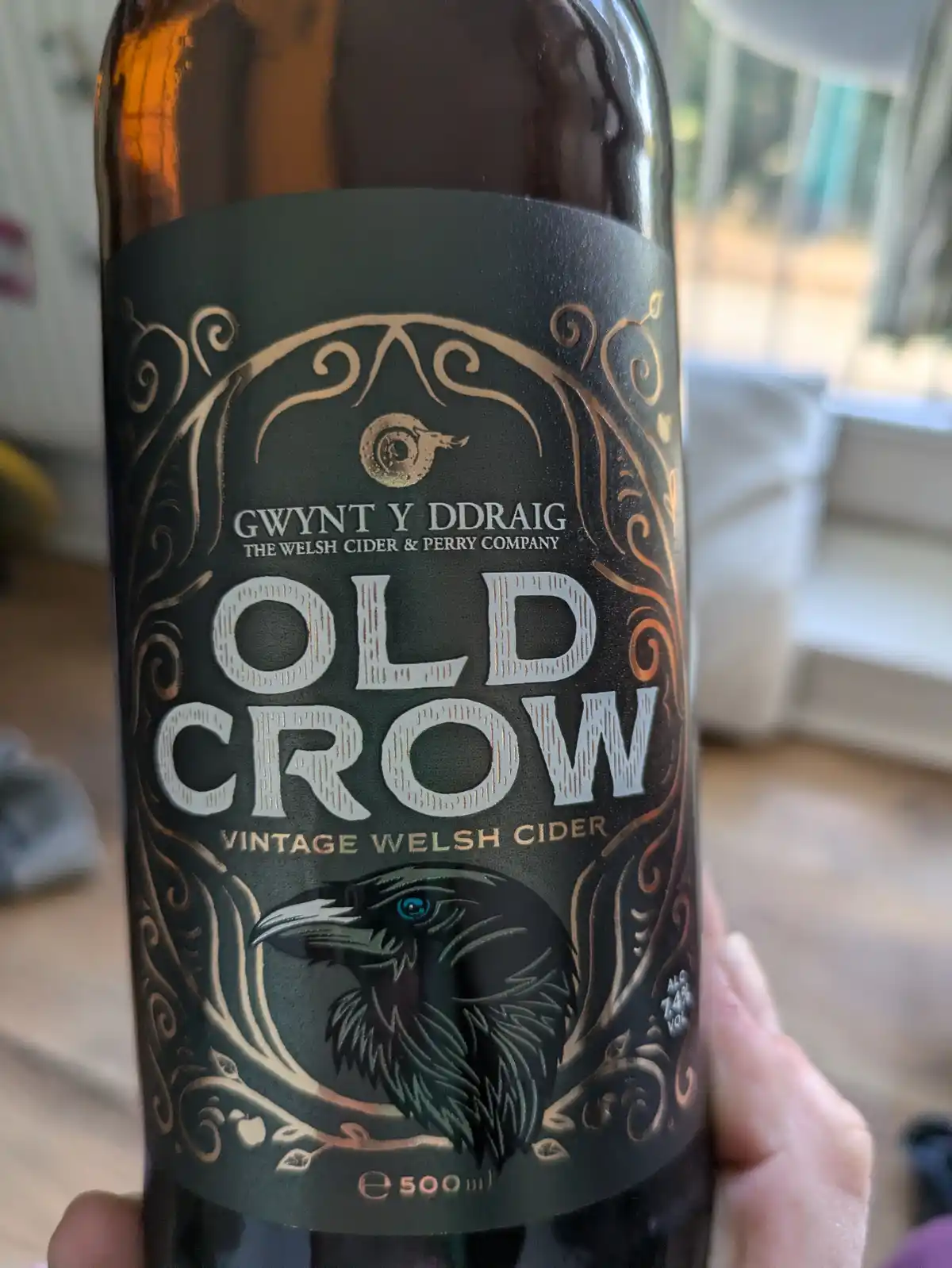 A close-up photo shows a hand holding a dark brown glass bottle of 'Old Crow Vintage Welsh Cider' by 'Gwynt Y Ddraig The…