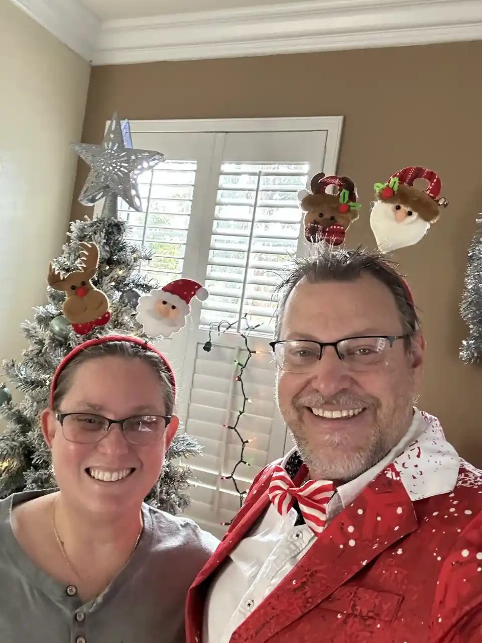 A smiling man and woman pose for a selfie in front of a decorated Christmas tree. Both wear festive headbands with plush…