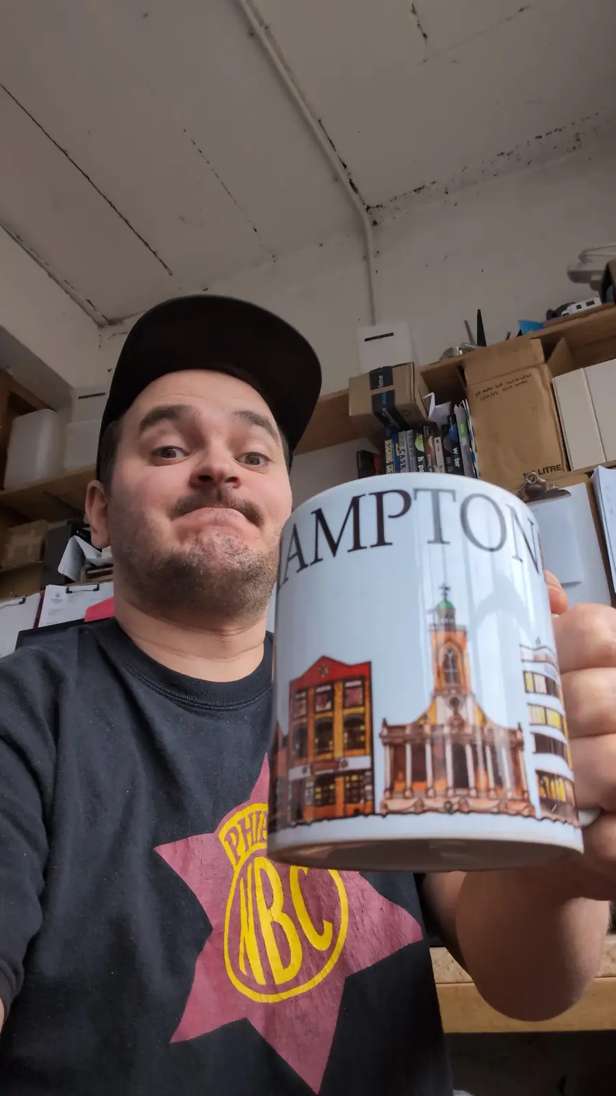 A man wearing a black cap and t-shirt holds up a white mug with 'HAMPTON' printed on it and an illustration of buildings. He…